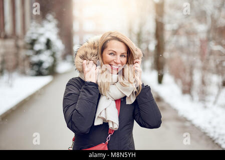 Gentile giovane donna bionda all aperto in inverno in piedi in una strada innevata cuddling giù nella sua calda sciarpa di lana e pelliccia cappuccio foderato di suo cappotto come lei Foto Stock