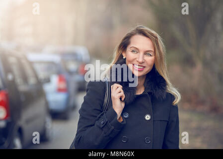 Elegante Attraente sorridente donna bionda in una nebbiosa strada in inverno indossando un cappotto caldo e borsetta sopra la sua spalla radente permanente a lato Foto Stock