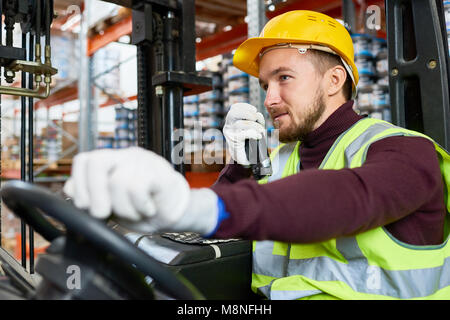 Vita Ritratto di giovane uomo seduto nel carrello e utilizzando un walkie-talkie durante la movimentazione merci in magazzino, spazio di copia Foto Stock