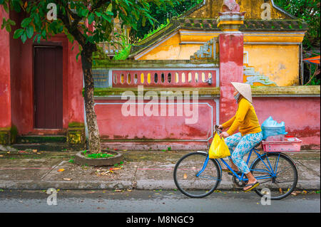 Bicicletta del Vietnam, vista di una donna che indossa un cappello conico in bicicletta passando attraverso le pareti colorate nel quartiere della città vecchia di Hoi An, Vietnam centrale. Foto Stock