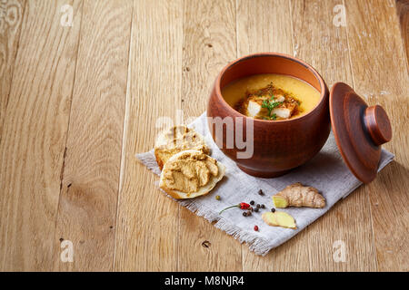 Pentola di creta di piccante cremosa zuppa di zucca guarnito con aneto, servita con crostini di baguette fette di pane ricoperte con pasta, lo zenzero su sfondo rustico, Foto Stock