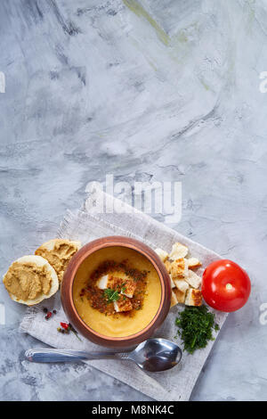 Pentola di creta di piccante cremosa zuppa di zucca guarnito con aneto, servita con pane tostato coperte con pasta e pomodoro rosso su bianco a trama, sfondo Foto Stock