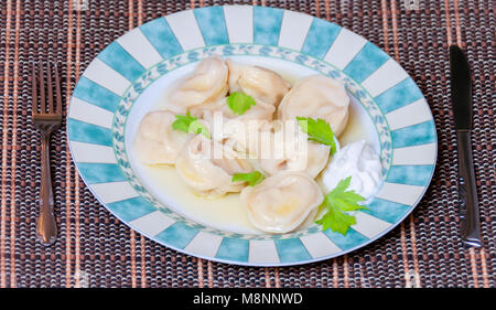 Appetitosi gnocchi bolliti con panna acida su un gran bel piatto con un fresco verde foglia, un coltello di metallo e la forcella giacciono sui bordi di un tappetino di rattan Foto Stock