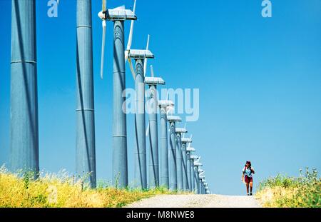 Donna che cammina sulla via accanto a turbine eoliche, parte del massiccio di wind farm complesso ad Altamont vicino Livermore, California, Stati Uniti d'America Foto Stock