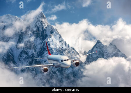 L'aereo è volare su nuvole basse contro le montagne con vette innevate in bright day. Paesaggio. Aereo passeggeri, cielo nuvoloso, rocce, neve. Il passeggero Foto Stock