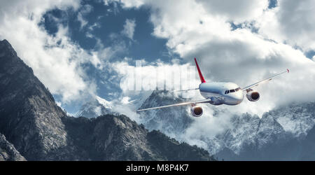 L'aereo è volare su nuvole basse contro le montagne con vette innevate in bright day. Paesaggio. Aereo passeggeri, cielo nuvoloso, rocce, neve. Il passeggero Foto Stock