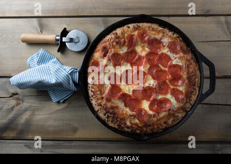 Deep Dish pizza in ghisa pan su tavola in legno rustico con pizza cutter e canovaccio avvolto attorno alla maniglia Foto Stock