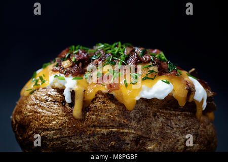Vista laterale di un carico di patate al forno con erba cipollina, bacon, formaggio cheddar, panna acida su nero Foto Stock