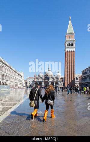 Due turisti in arancio brillante su scarponi a piedi attraverso Acqua Alta o alta marea allagamento, in Piazza San Marco, Venezia, Veneto, Italia come l'acqua r Foto Stock