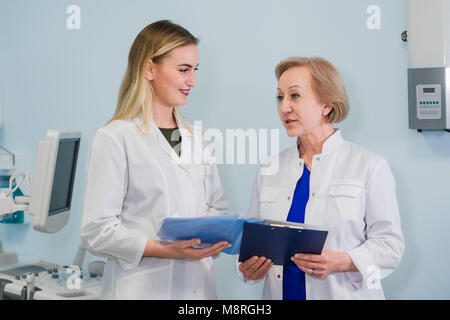 Senior medico parlando con la giovane donna in piedi assistente in ufficio ginecologiche con sedia e lampada sullo sfondo Foto Stock