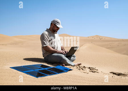 Uomo che lavora con un laptop che si ricarica da un pannello solare portatile all'aperto nel deserto. Lavorare in remoto con gadget, autosufficienza e cielo blu ecocompatibile Foto Stock