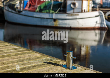 Messa a fuoco poco profonda su chrome bollard sul molo in legno bianco con la barca da pesca sfumata in background. Foto Stock