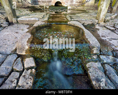 Acqua fresca che scorre da un pozzetto a Tissington villaggio con donazioni di denaro per desideri nel Parco Nazionale di Peak District, Derbyshire, England, Regno Unito Foto Stock