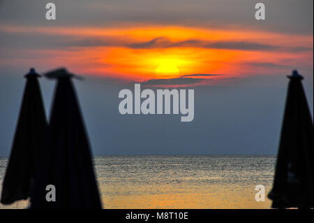 Tramonto tropicale al cielo, con il sole che tramonta dietro le nuvole in una gloriosa bagliore arancione. Ocean scena inquadrata da ombrelloni da spiaggia. Prese a Kata Noi, Thailandia Foto Stock