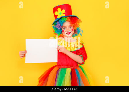 Funny bambina in costume da clown azienda tavolo bianco per voi la pubblicità. Studio shot, isolato su sfondo giallo Foto Stock