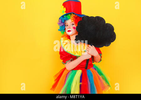 Infelice fanciulla in costume da clown e cappello nero di contenimento tabella nella forma di una nuvola. Studio shot, isolato su sfondo giallo Foto Stock