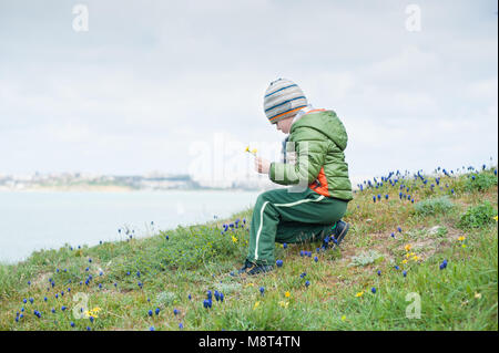 Bello piccolo ragazzo con il tarassaco giallo in mano on Grassy Slope fioritura con Muscari fiori sullo sfondo del mare in primavera Foto Stock