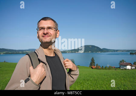 Un turista con lo zaino escursionismo su una strada di campagna vicino al lago. Una piccola città e il verde delle colline in background Foto Stock