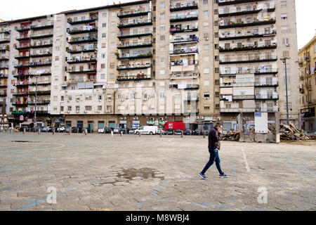 Un uomo sul suo telefono cellulare passeggiate attraverso Piazza Mercato a Napoli, Italia, con un casamento blocco di appartamenti come sfondo Foto Stock