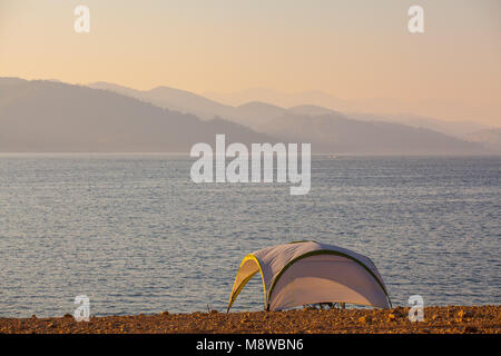 Piccolo Rifugio sulle rive del lago di montagna con sagome delle montagne sull'orizzonte al tramonto Foto Stock