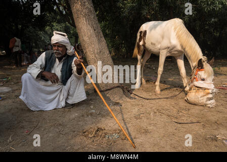 Un uomo si siede sul suo haunches mentre il suo cavallo alimenta in Sonepur Mela, Sonepur, India. Foto Stock
