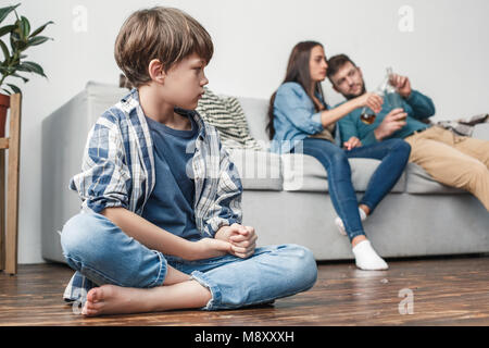 Madre padre e figlio problemi sociali alcolismo piccolo ragazzo seduto sul pavimento vicino guardando i genitori a bere birra infelice Foto Stock
