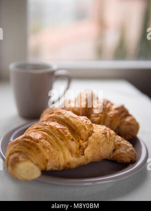 Vista ravvicinata dei due croissant francesi con una tazza di una bevanda calda nella parte anteriore di una finestra in un giorno di pioggia Foto Stock