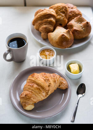Vista al di sopra di una prima colazione continentale con croissant, burro, marmellata, caffè nero e un cesto di un assortimento di prodotti di pasticceria francese sul tavolo bianco Foto Stock