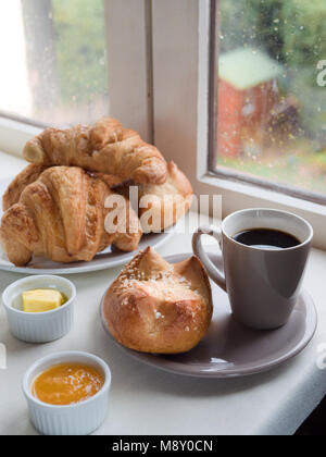 Al di sopra di vista di una brioche, Francese bun , croissant, con burro e marmellata e una tazza di caffè davanti a una finestra in un giorno di pioggia Foto Stock