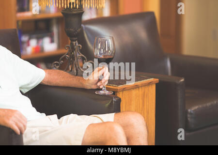 Un uomo maturo con in mano un bicchiere di vino rosso mentre vi rilassate in un cuoio arnchair Foto Stock