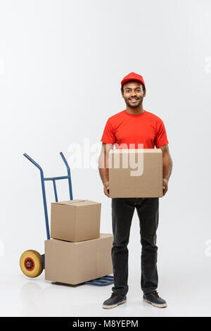 Concetto di consegna - Ritratto di bello americano africano uomo di consegna o corriere con carrello a mano e della scatola di contenimento. Isolato su grigio di sfondo per studio. Copia dello spazio. Foto Stock