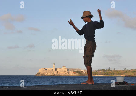 Un ragazzo di pesci a mano dalla seawall di Malecon a l'Avana, Cuba, con la fortezza nota come Morro Castle (El Morro) in background. Foto Stock