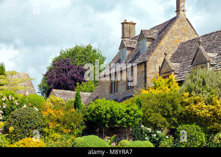 Appartata di pietra calcarea Cotswold House con tetto in ardesia, nel paese di lingua inglese giardino pieno di piante sempreverdi, alberi, fiori, rosa rose, su di un soleggiato estate Foto Stock