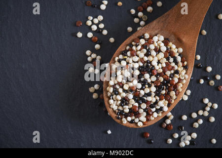 Materie miste di quinoa (Chenopodium quinoa). Rosso, bianco e nero semi in cucchiaio di legno su sfondo nero. Vista da sopra con lo spazio di copia Foto Stock