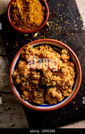 Angolo di alta vista di una casseruola di terracotta con il couscous con il pollo e le verdure su una tavola in legno rustico, accanto a una ciotola con il curry in polvere Foto Stock