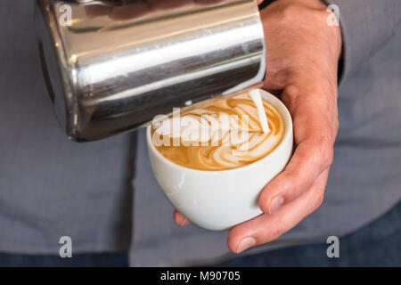 Professional barista versando latte vaporizzato da acciaio inossidabile lo sbarramento rotante nella tazza da caffè rendendo latte art sul cappuccino Foto Stock