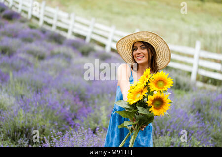 Giovane donna tenendo un mazzo di fiori di Sun nel mezzo di un campo di lavanda, indossa un elegante vestito blu e un cappello di paglia. Foto Stock