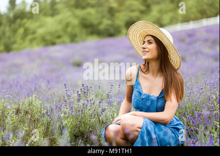 La donna al centro di un campo di lavanda, indossa un elegante vestito blu e un cappello di paglia, posa con un grande sorriso. Foto Stock