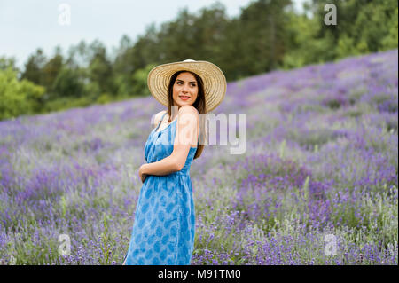 La donna al centro di un campo di lavanda, indossa un elegante vestito blu e un cappello di paglia, posa con un grande sorriso. Foto Stock