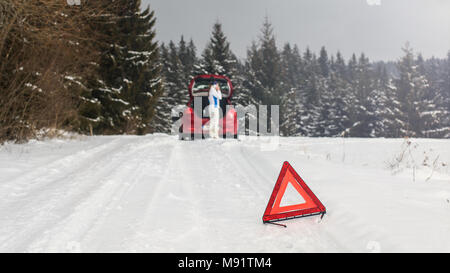 Brillante triangolo rosso su una strada innevata avvertenza altri piloti con la donna in piedi accanto a Broken auto in foresta, chiamando il servizio di riparazione sul suo telefono cellulare Foto Stock