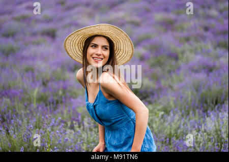 La donna al centro di un campo di lavanda, indossa un elegante vestito blu e un cappello di paglia, posa con un grande sorriso. Foto Stock