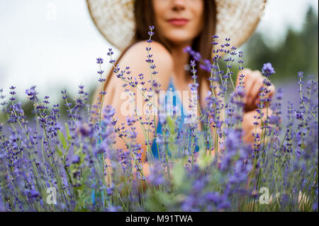 Close-up di fiori di lavanda con una giovane e bella donna su sfondo indossando un cappello di paglia in piedi in un grande campo di piante di lavanda Foto Stock