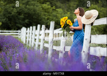 Giovane donna tenendo un mazzo di fiori di Sun nel mezzo di un campo di lavanda, indossa un elegante vestito blu e un cappello di paglia. Foto Stock