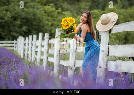 Giovane donna tenendo un mazzo di fiori di Sun nel mezzo di un campo di lavanda, indossa un elegante vestito blu e un cappello di paglia. Foto Stock