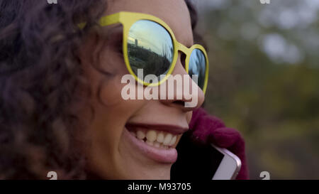 Primo piano della biracial felice lady in occhiali da sole parlando al telefono, riflessione della città Foto Stock