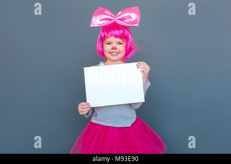 Funny bambina in costume da clown holding scrivania bianca. Studio shot, isolata su uno sfondo grigio Foto Stock