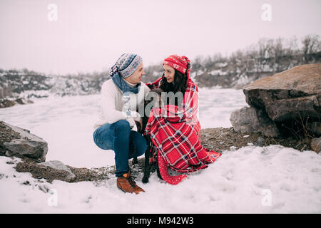 Coppia giovane in cappelli e sciarpe con il cane avvolto in rosso plaid durante l'inverno a piedi accanto al lago ghiacciato e rocce. Foto Stock