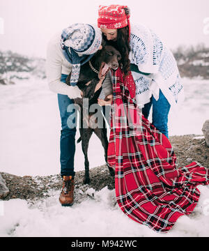 Coppia giovane in cappelli e sciarpe avvolto in rosso plaid sono in piedi e il bacio di cane accanto al lago ghiacciato e rocce durante l'inverno a piedi. Foto Stock