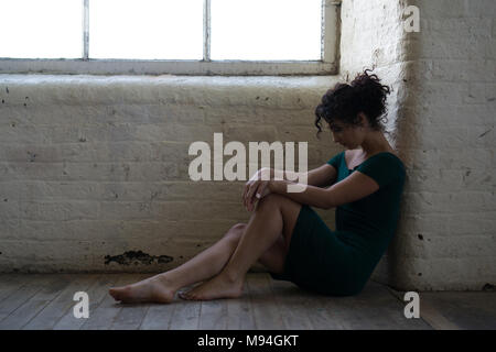 Vista laterale di un triste giovane donna che indossa un abito sabato sul pavimento mediante la finestra Foto Stock