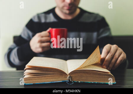 Uomo in scuro maglione a strisce seduti a un tavolo di legno e la lettura prenota tenendo la tazza. Serata di routine per la lettura di libri e una tazza di tè nel anche Foto Stock